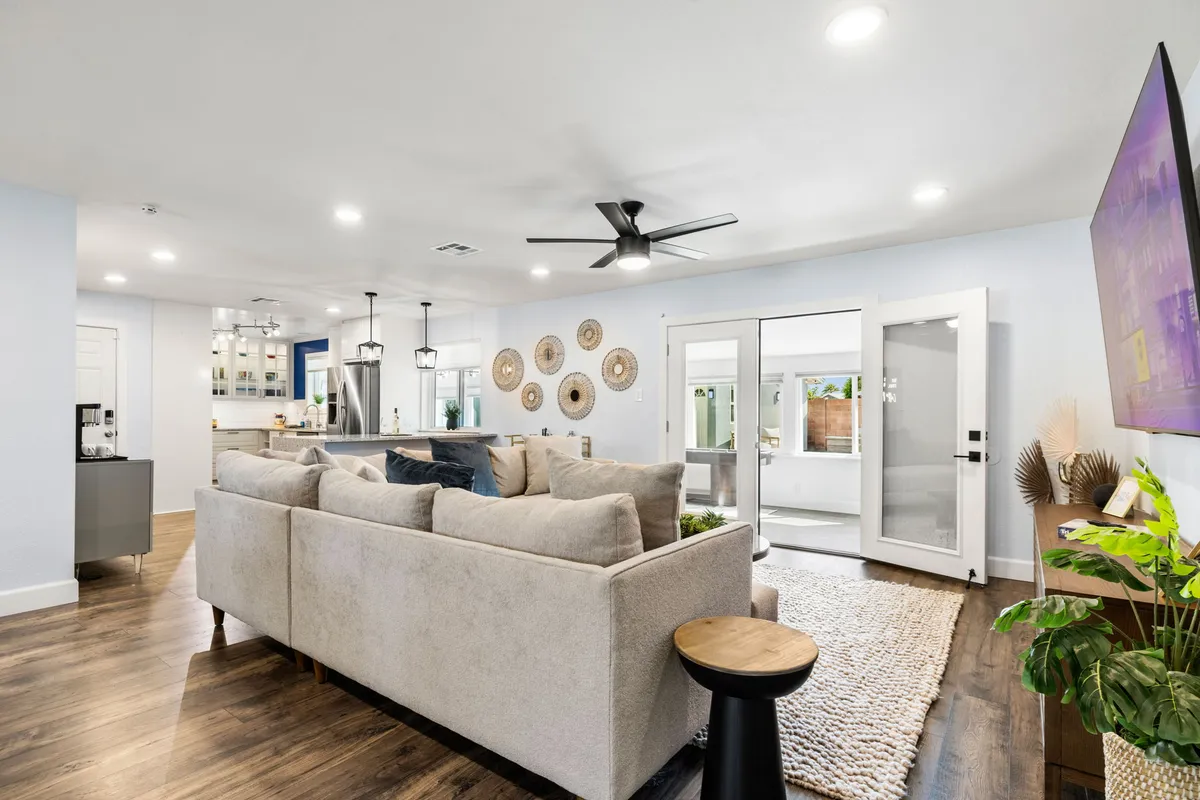 This image features a plush sectional sofa, decorative wall plates, open doors that lead outside, a stylish ceiling fan, and a TV mounted on the wall. The space is arranged for comfortable lounging and group conversations, with plenty of seating and a welcoming layout.