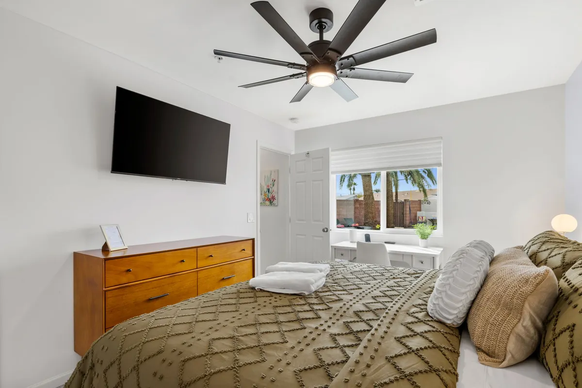 A tranquil bedroom with a bed featuring decorative pillows, a wooden dresser, a wall-mounted TV, and a window with a view. The space feels peaceful and offers both comfort and modern entertainment options.