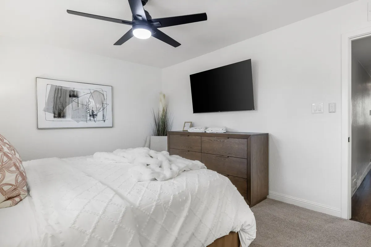 A comfortable bedroom setup with a plush bed covered in a white quilted coverlet, a large flat-screen TV mounted on the wall, a wooden dresser, and contemporary abstract wall art. The setting is relaxed and ideal for watching TV or reading in bed.
