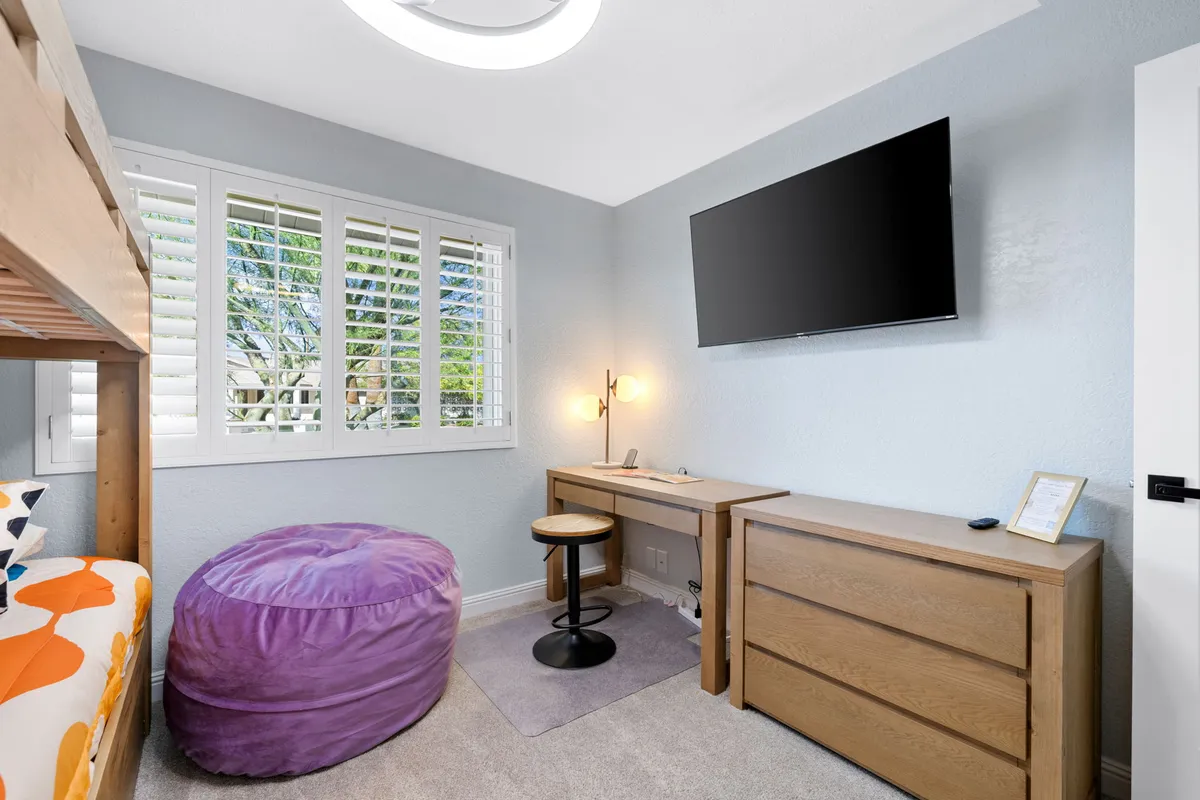 A playful and inviting bedroom featuring a bunk bed with colorful bedding, a purple bean bag chair, a wooden desk and chair, a television, and a window with shutters. The space is designed for both leisure and activity, appealing to families and groups.