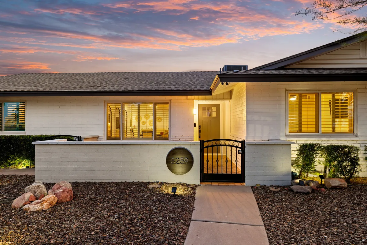 This exterior image displays the house's welcoming entrance gate, the illuminated facade, and thoughtfully designed landscaping, all under a sunset sky. The warm lights glowing from the windows hint at inviting interiors and active social spaces within.