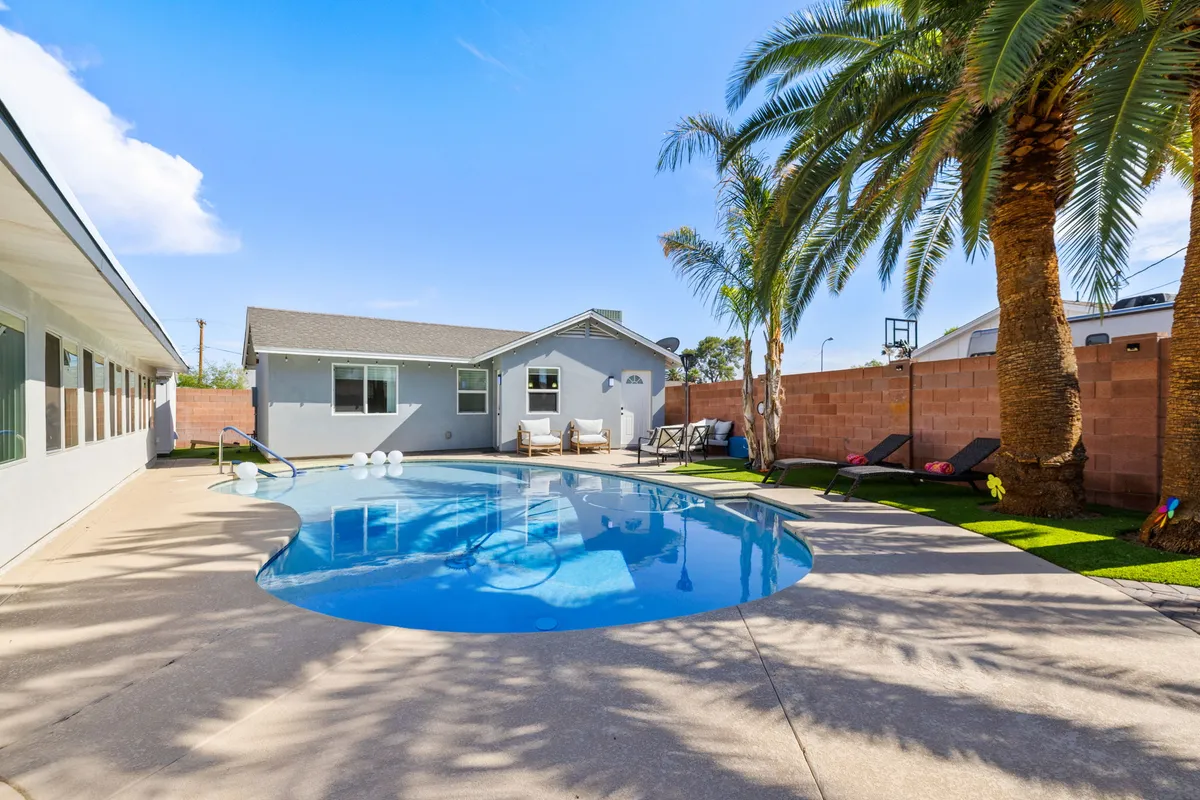 The image showcases a sparkling swimming pool surrounded by towering palm trees, plush lounge chairs, and a well-maintained backyard wall, all bathed in warm Arizona sunlight. The inviting outdoor space hints at relaxing poolside afternoons, sunset gatherings, and vibrant group activities like basketball and barbecues.