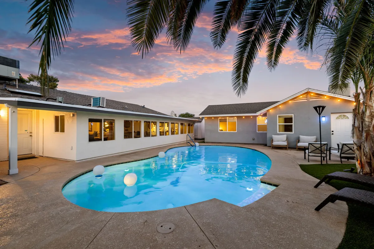 This exterior image captures a sparkling swimming pool surrounded by palm trees and inviting outdoor seating, with twinkling lights illuminating the house as dusk falls. The lively ambiance and festive setting make it perfect for evening gatherings and celebrations.