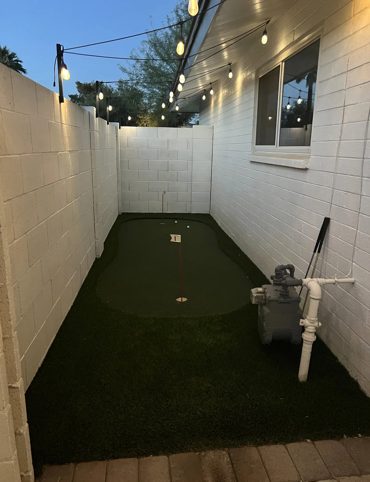 An inviting backyard putting green illuminated with string lights, bordered by white walls and surrounded by cozy gathering spaces. The scene is set during twilight, creating a warm and festive ambiance perfect for both golf practice and casual socializing.