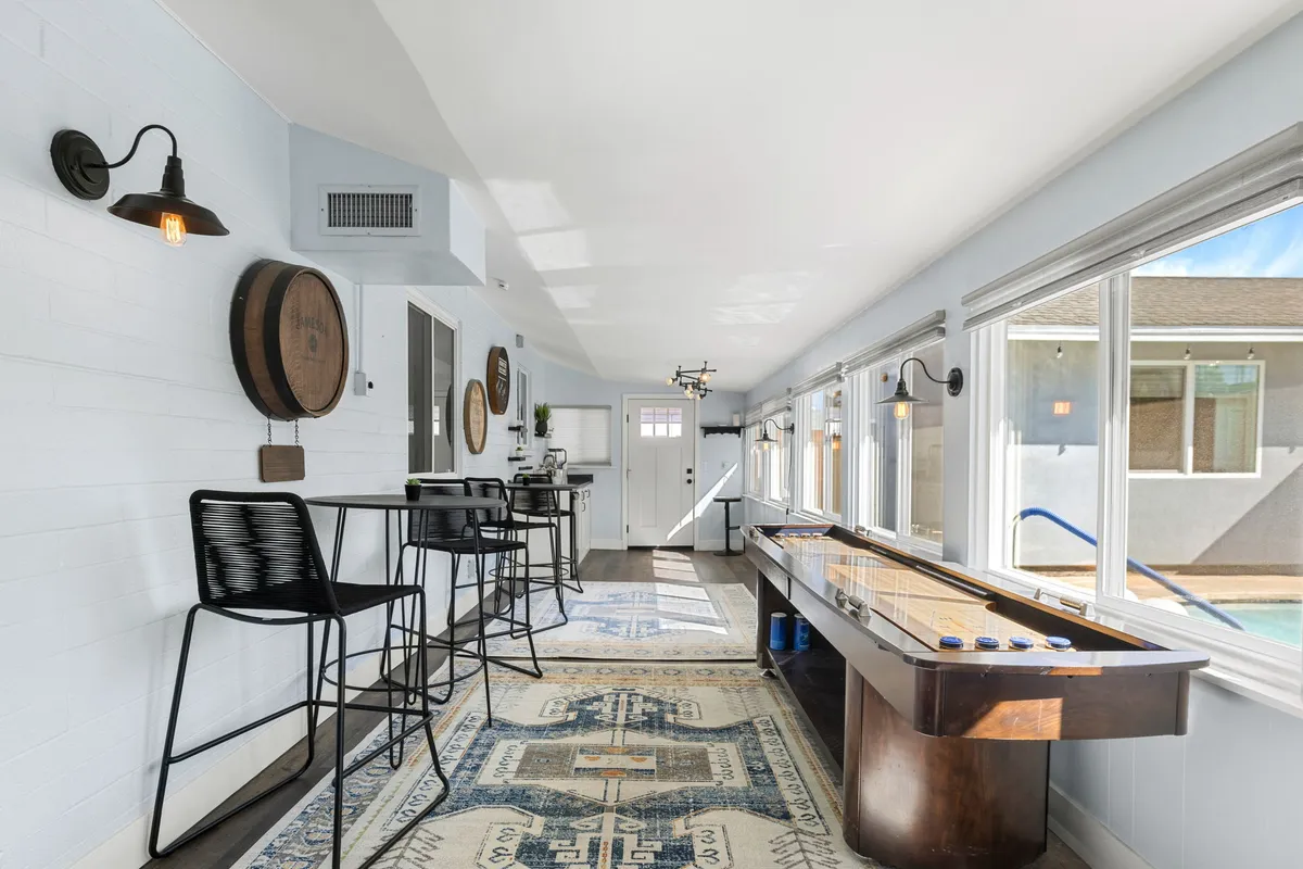 A stylish game room interior with a shuffleboard table, barrel wall decor, and bar stools. Large windows let in natural light, creating a cheerful, casual vibe ideal for socializing and friendly competition.