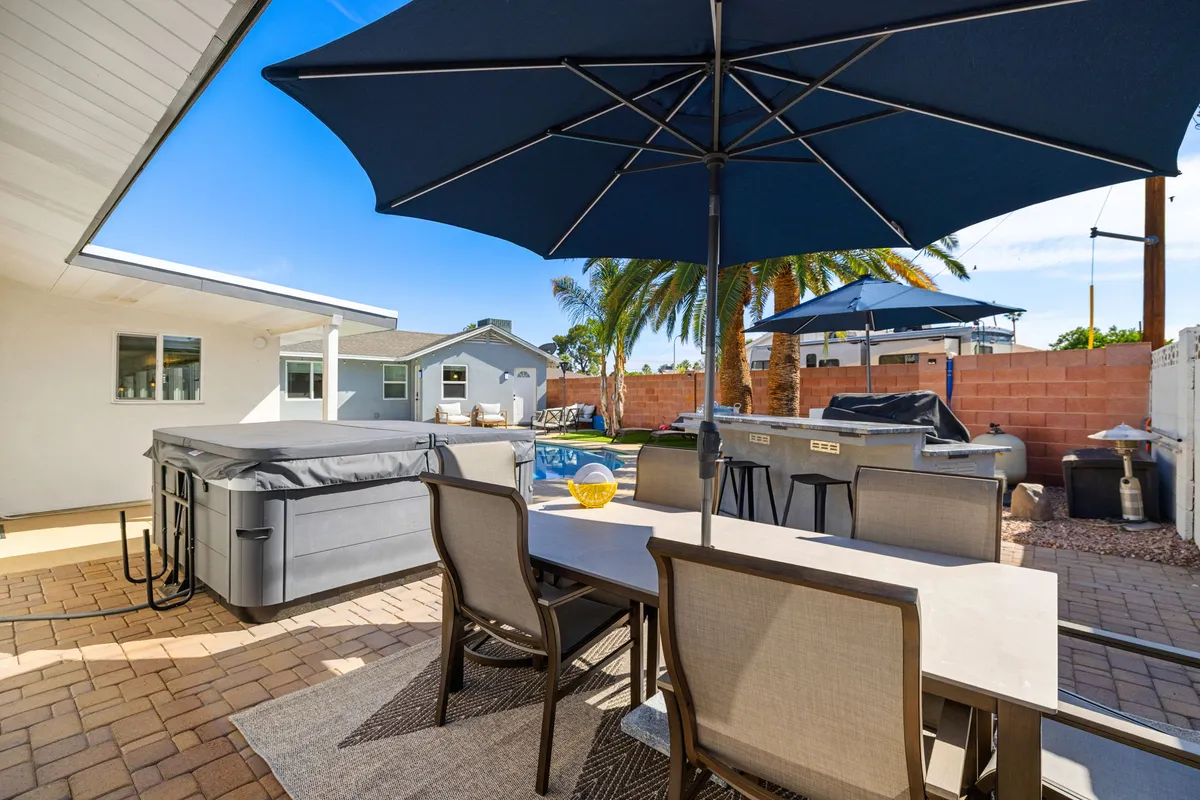 This exterior shot features a relaxing hot tub, an alfresco dining table, umbrellas, and lush palm trees, all bathed in soft outdoor lighting. The scene evokes the idea of coming home after a night out to continue socializing or relax in luxury.